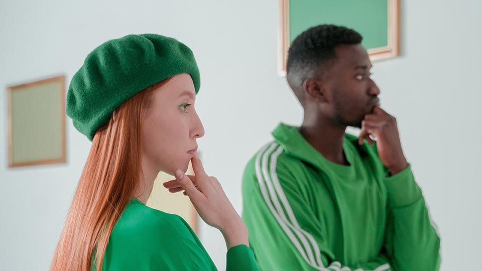Two people wearing all green ponder framed images on a wall.
